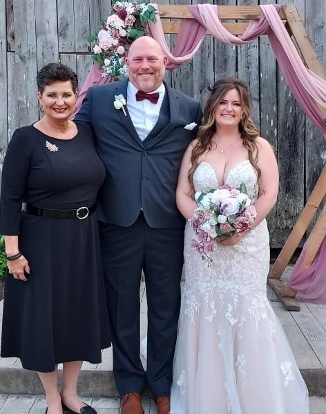 Three people posing at a wedding, with the bride holding a bouquet in a white gown and pink drapes behind them.