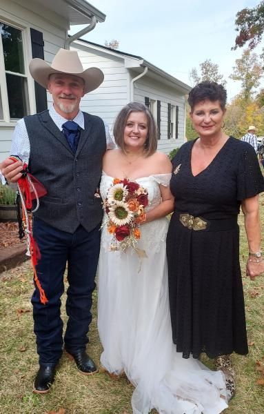 Wedding couple standing outdoors, bride in white dress holding bouquet, beside man in cowboy hat and woman in black dress
