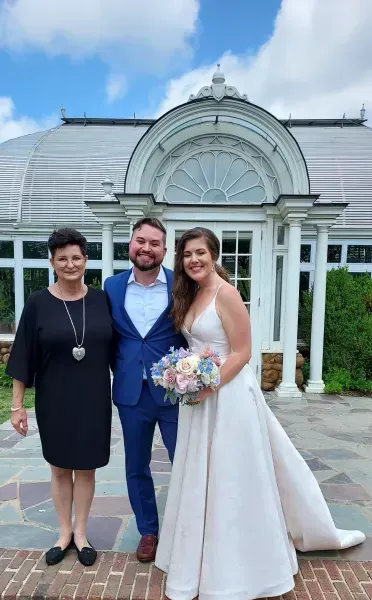 Three people pose smiling in front of a glass conservatory, with one wearing a white wedding dress.