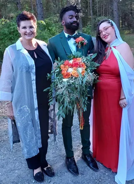 Three people posing outdoors, with the center person holding a large floral arrangement.