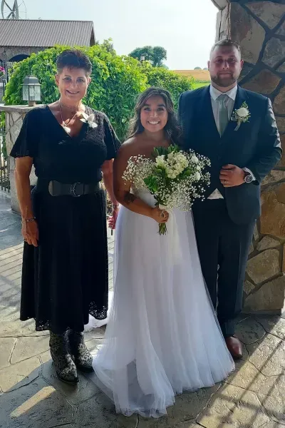 Three people posing outdoors at a wedding, with the bride holding a bouquet in a white dress.
