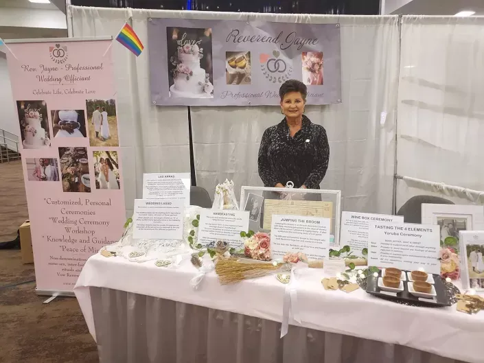 Woman at a memorial display booth with photos, flowers, candles, and handwritten signs behind a white table.