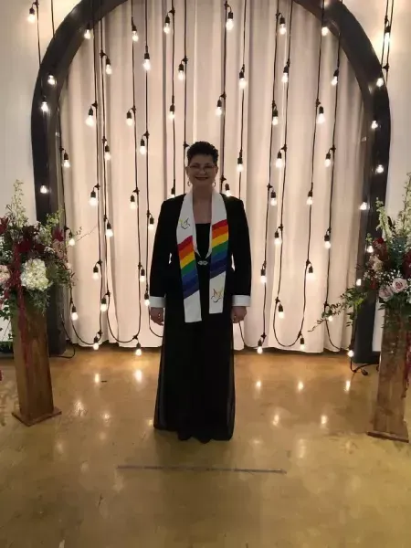 Person standing in a black gown with a rainbow stole before a lit draped backdrop and floral arrangements