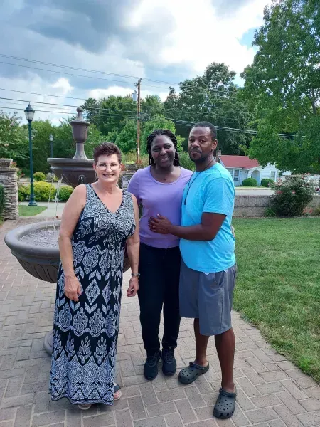 Three people smiling on a brick path in a park, with trees and grass in the background.