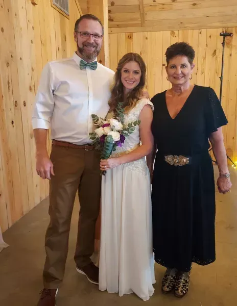 Three people smiling in a wooden room, with the center woman holding a bouquet and wearing a white dress.