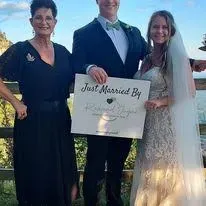Three people at an outdoor wedding hold a “Just Married” sign as the bride and groom smile.