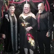 Three people posing together in black outfits, one in a black dress with gold floral accents.