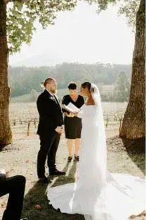 Outdoor wedding ceremony by a lake, with an officiant reading vows beside the bride and groom.