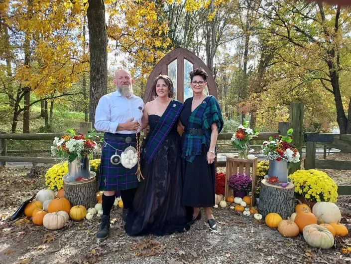 Three people in dark formal outfits standing among pumpkins and autumn flowers outdoors