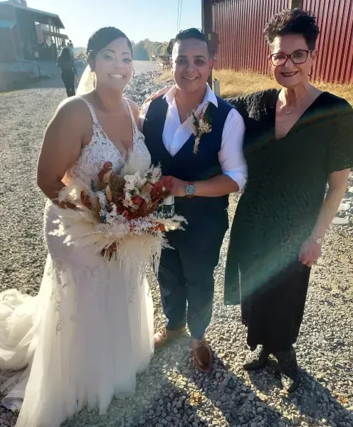 Three people smiling outdoors, with one in a white dress holding a bouquet at a rustic wedding.