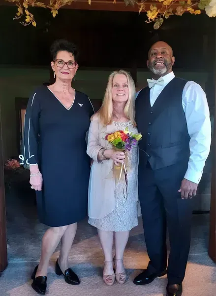 Three people posing indoors, with the center person holding a bouquet of flowers.