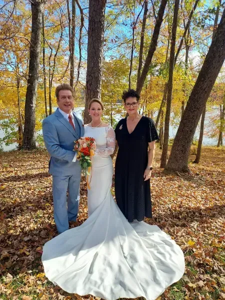 Three people posing outdoors in autumn; one wears a white wedding gown and holds a bouquet.
