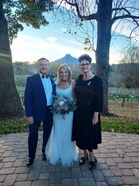 Three people standing outdoors at a wedding, with the bride holding a bouquet and the groom in a suit.