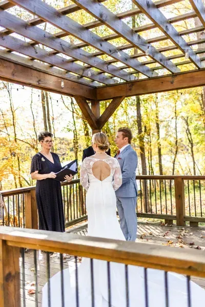 Wedding ceremony under a wooden pavilion in autumn, with an officiant and couple facing each other.