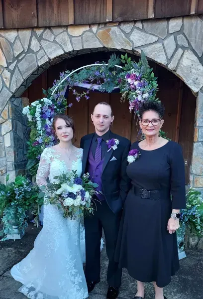 Bride and groom with a woman in black dress posing under a stone arch with purple flowers
