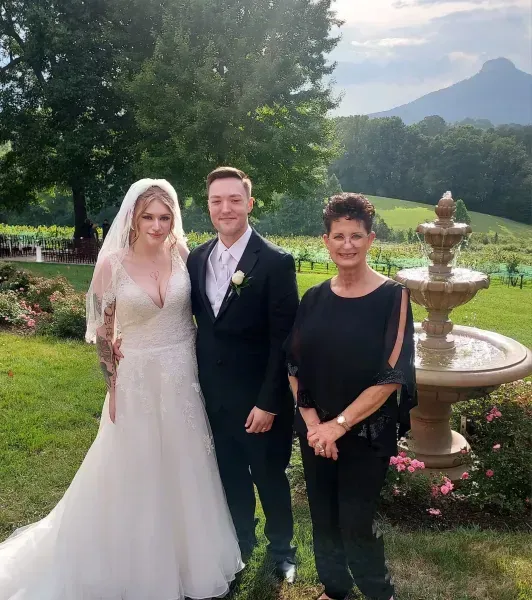 Bride, groom, and guest pose in a garden beside a fountain with mountains in the background.