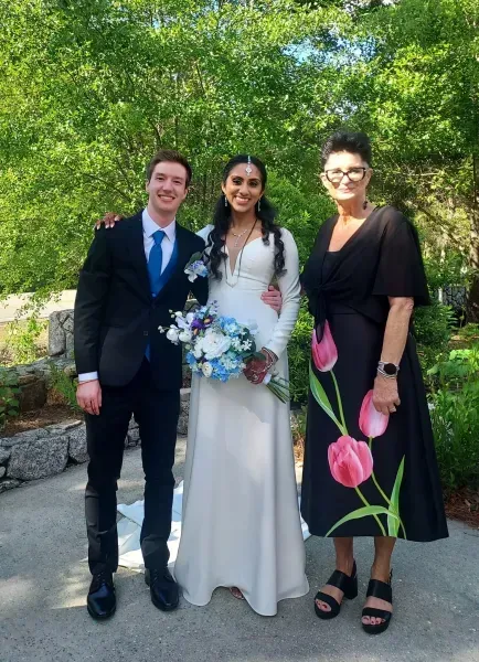 Three people pose outdoors in formal attire, with a bride holding a blue and white bouquet.