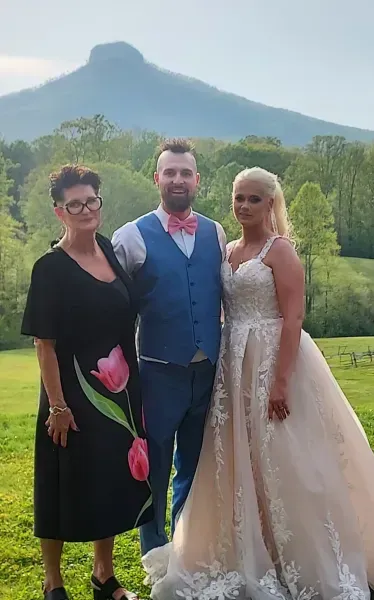 Three people pose in wedding attire outdoors with a mountain backdrop.