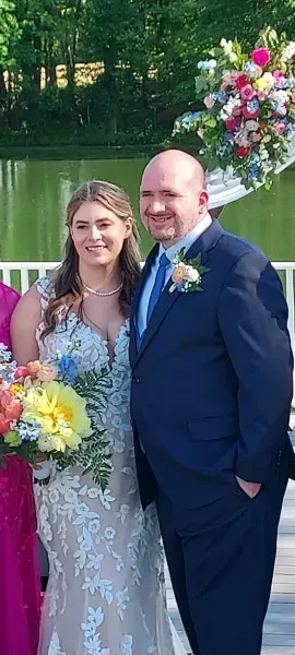 Couple posing in formal attire beside a lake, holding a colorful bouquet and floral arrangement.