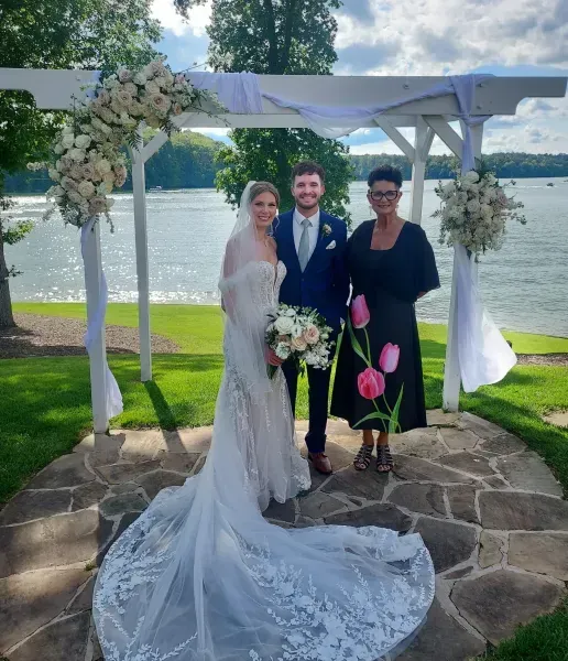 Bride and groom with officiant under floral arch by a lake on a sunny day