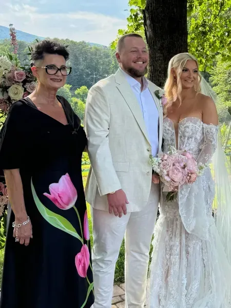 Three people posing outdoors at a wedding, with the bride in a white lace gown holding pink flowers.