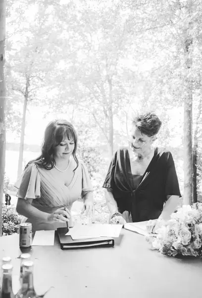 Two people seated outdoors at a table with flowers and an open book, one writing while the other looks on.