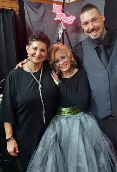 Three people smiling in formal attire at an event, standing before a dark curtain and pink neon sign