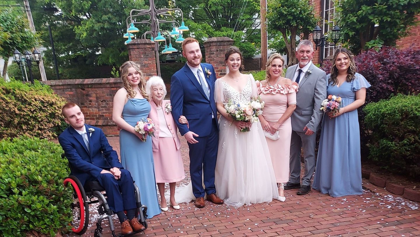 Wedding party posing outdoors in formal attire, with a bride holding a bouquet and a man in a wheelchair on the left.