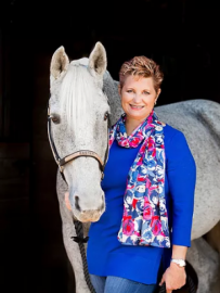 Woman in blue top and floral scarf standing beside a white horse against a black background