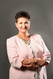 Woman standing with arms crossed, wearing a light pink blazer and necklace against a gray background