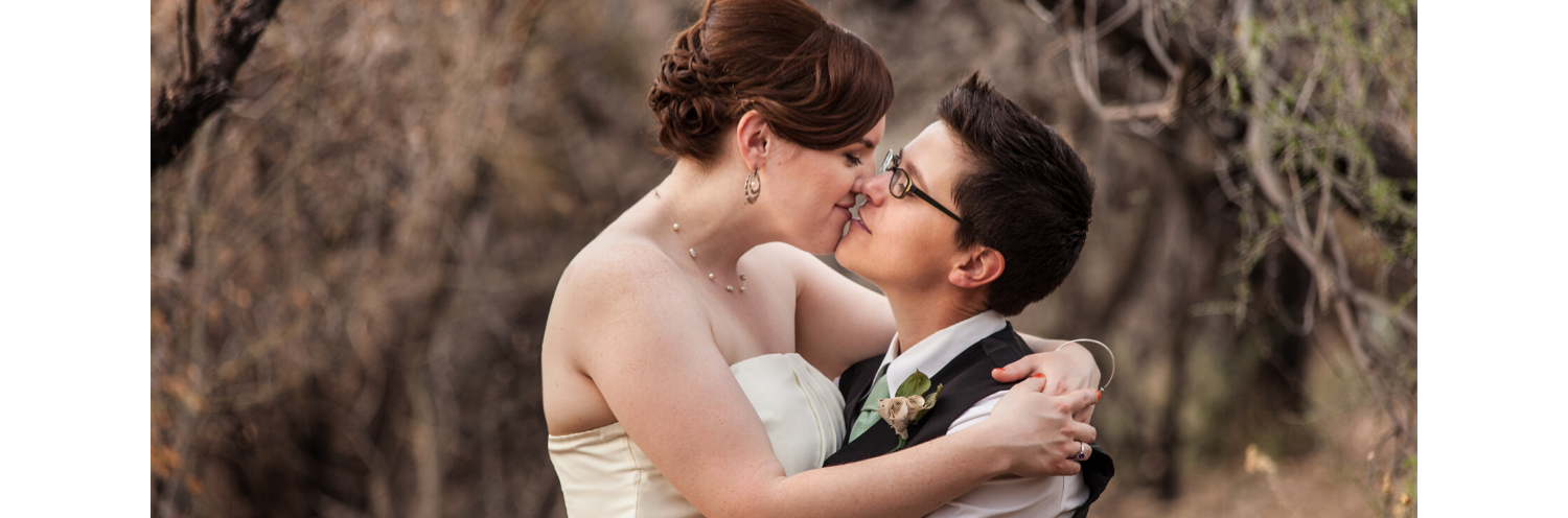 Couple kissing outdoors, bride in white dress and groom in glasses and dark suit, holding each other closely