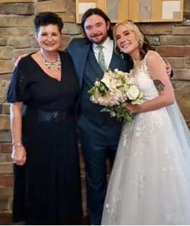 Three people posing at a wedding, with the bride in a white gown holding a bouquet beside two smiling guests.