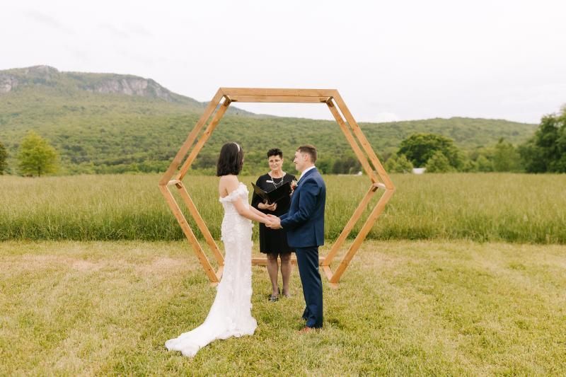 Wedding couple standing under a wooden hexagonal arch in a grassy field with mountains in the background