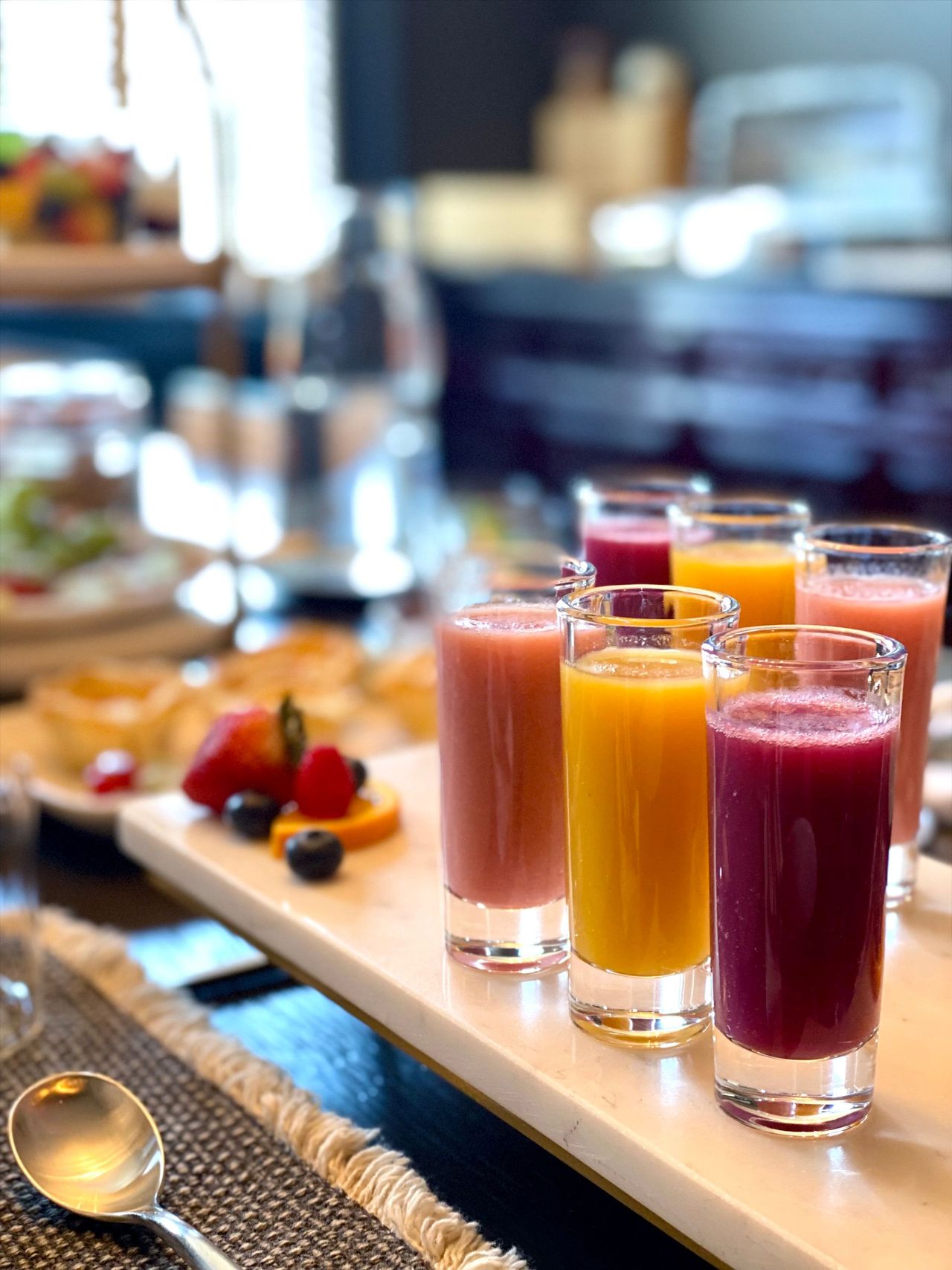 Assorted colorful juice shots on a buffet table, with spoons and fruit in the background