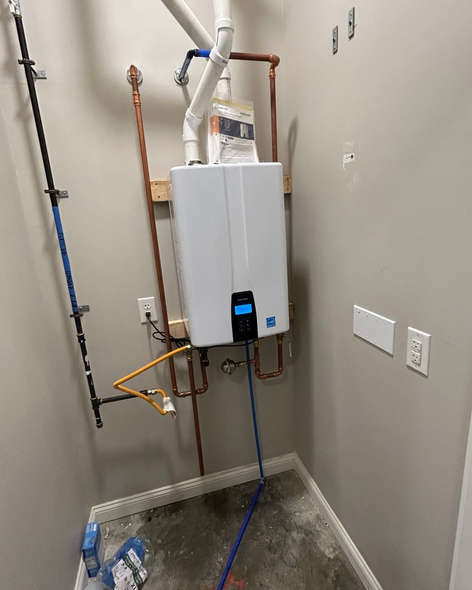A white tankless water heater mounted on a wall in a utility room with copper and blue pipes.