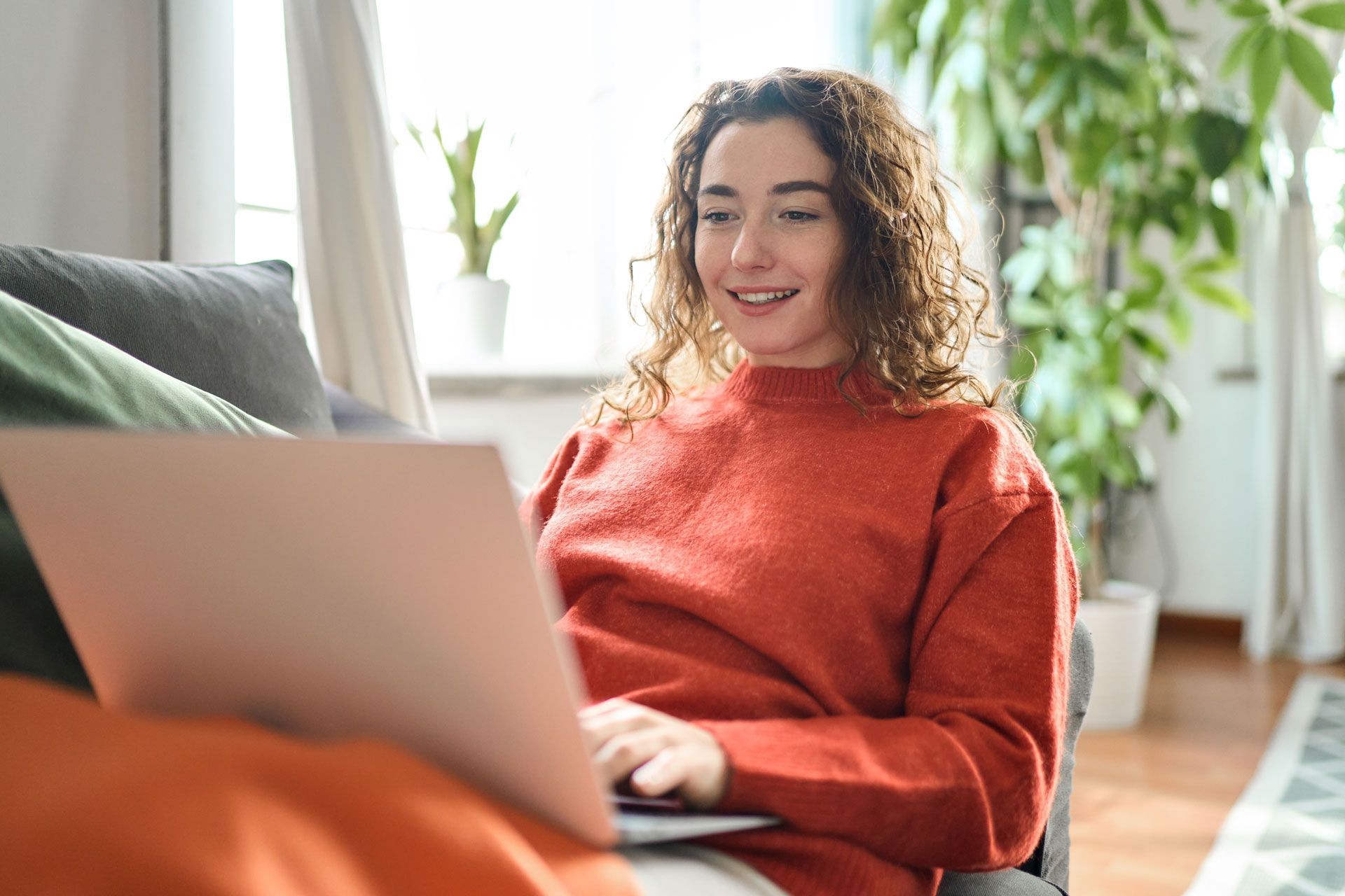 Woman smiling while using a laptop on a couch in a bright living room