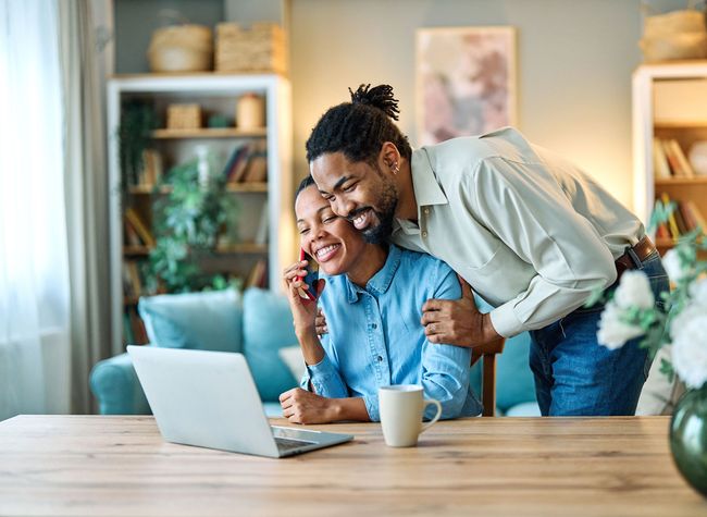 Two people smiling at a laptop in a cozy home office, with coffee cups on the table.