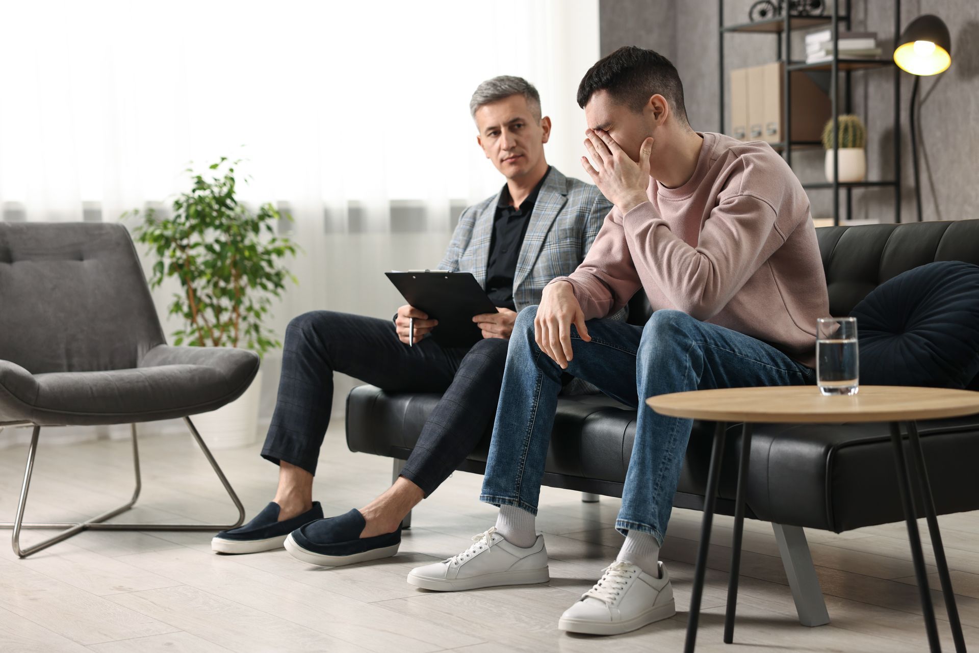 Therapist with a clipboard listens to a distressed person on a couch. The person covers their face.