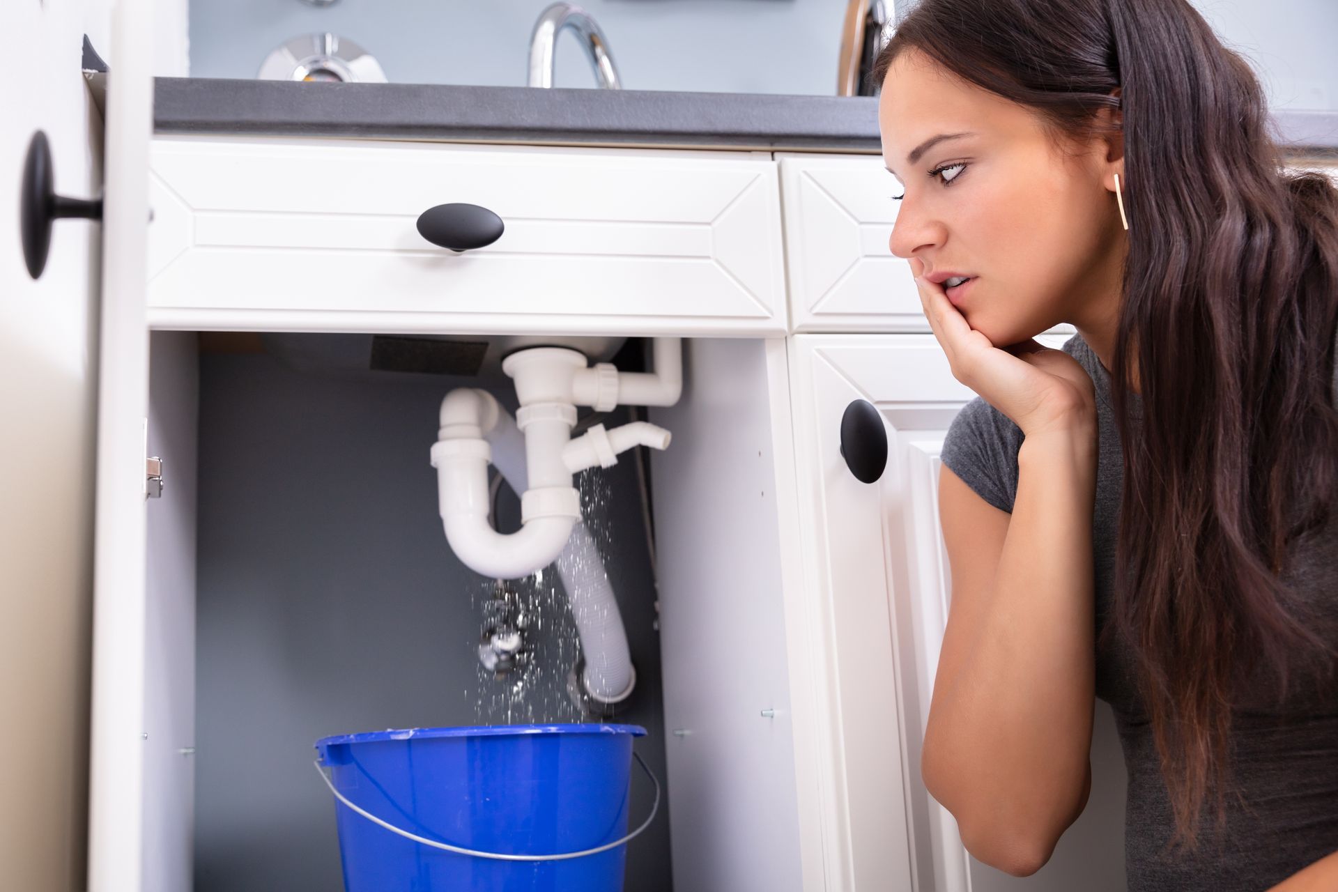 Woman looking under a sink at a leaking pipe into a blue bucket