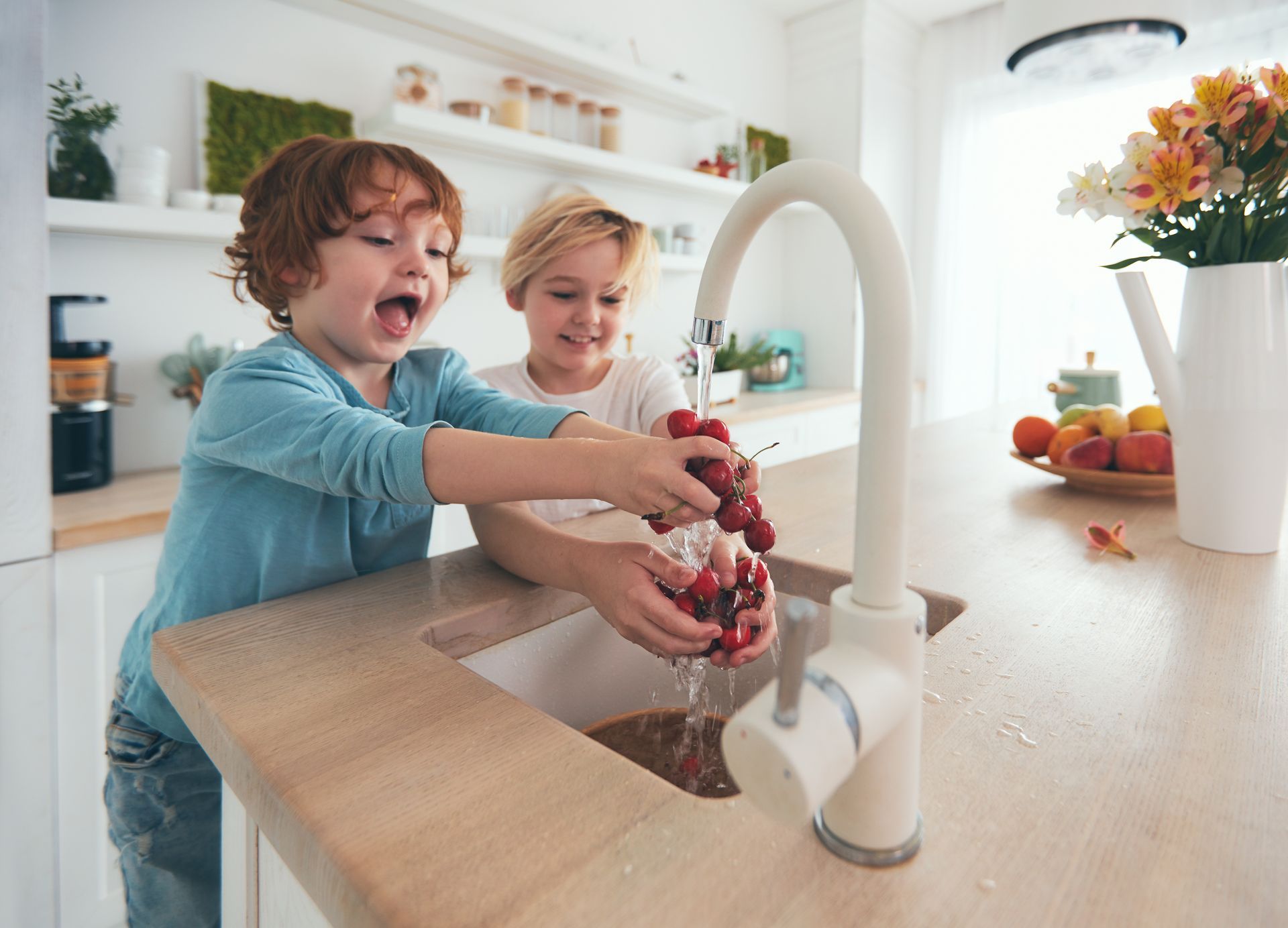 Two children washing red berries together at a kitchen sink in a bright home kitchen