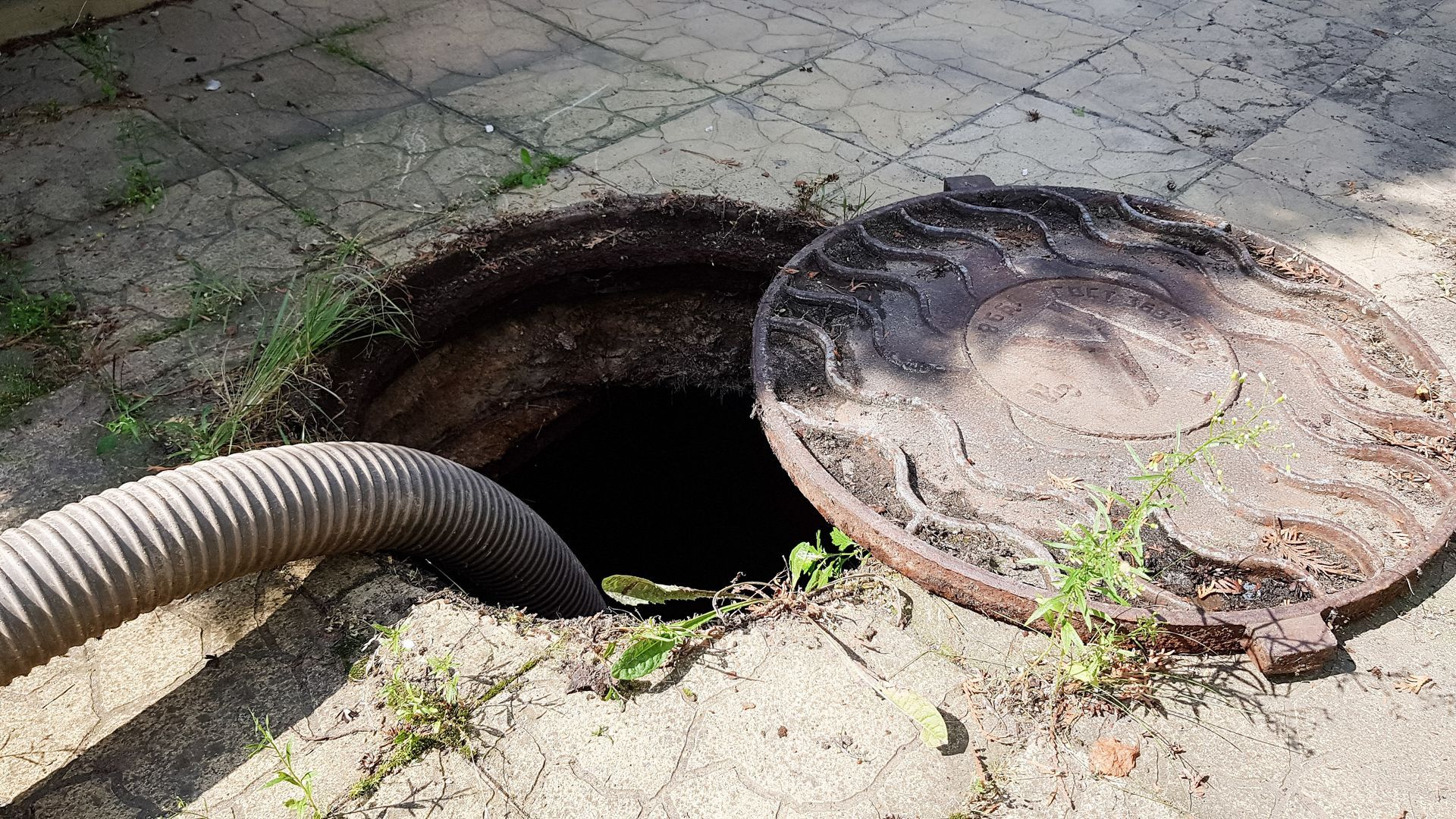 Open manhole with a hose leading into it on a paved surface, cover lying beside the opening