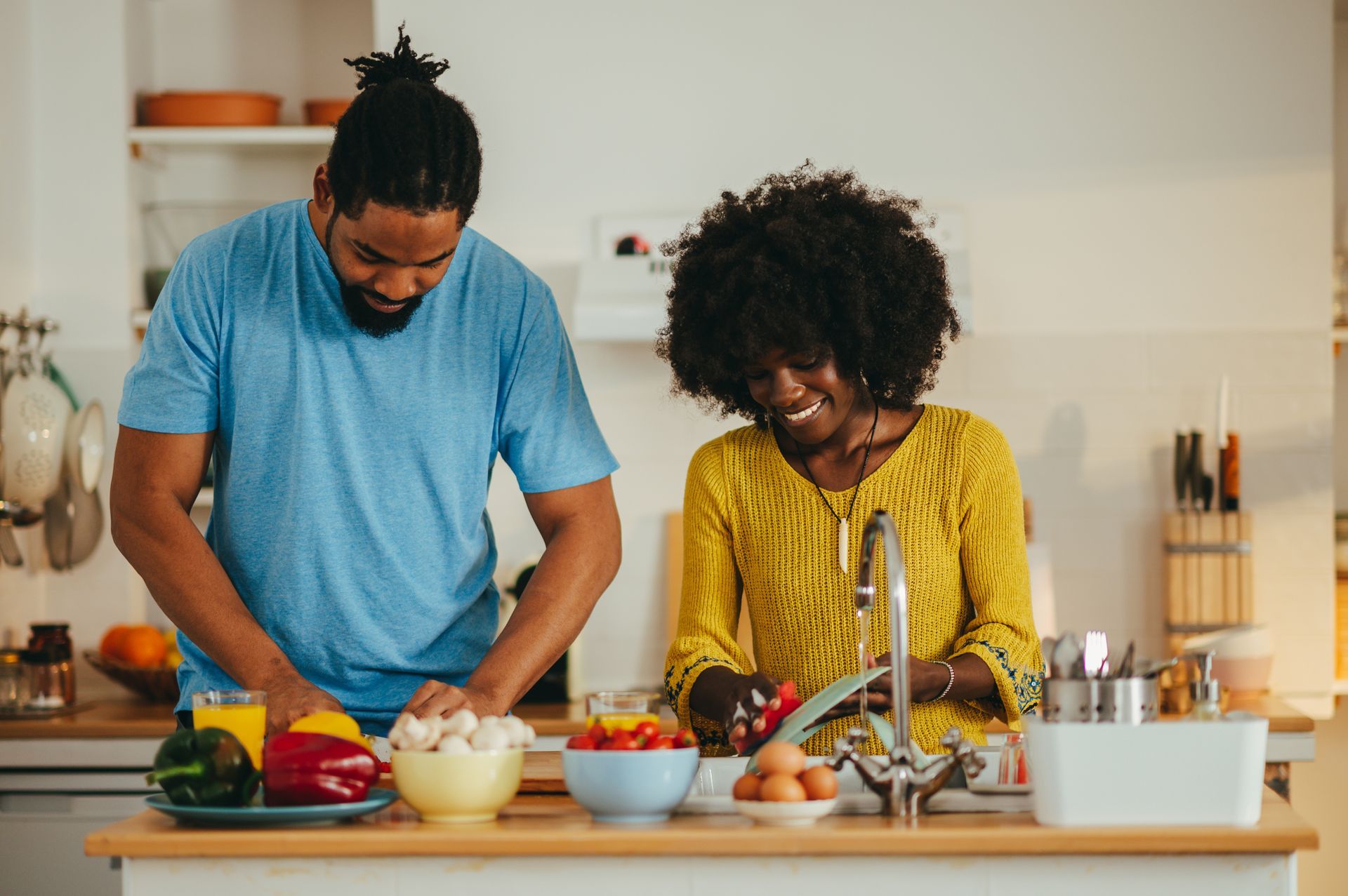 Two people preparing vegetables together in a bright kitchen, with bowls, fruit, and a sink on the counter