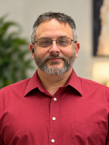Man in a red shirt and glasses, smiling in front of a softly blurred indoor background.