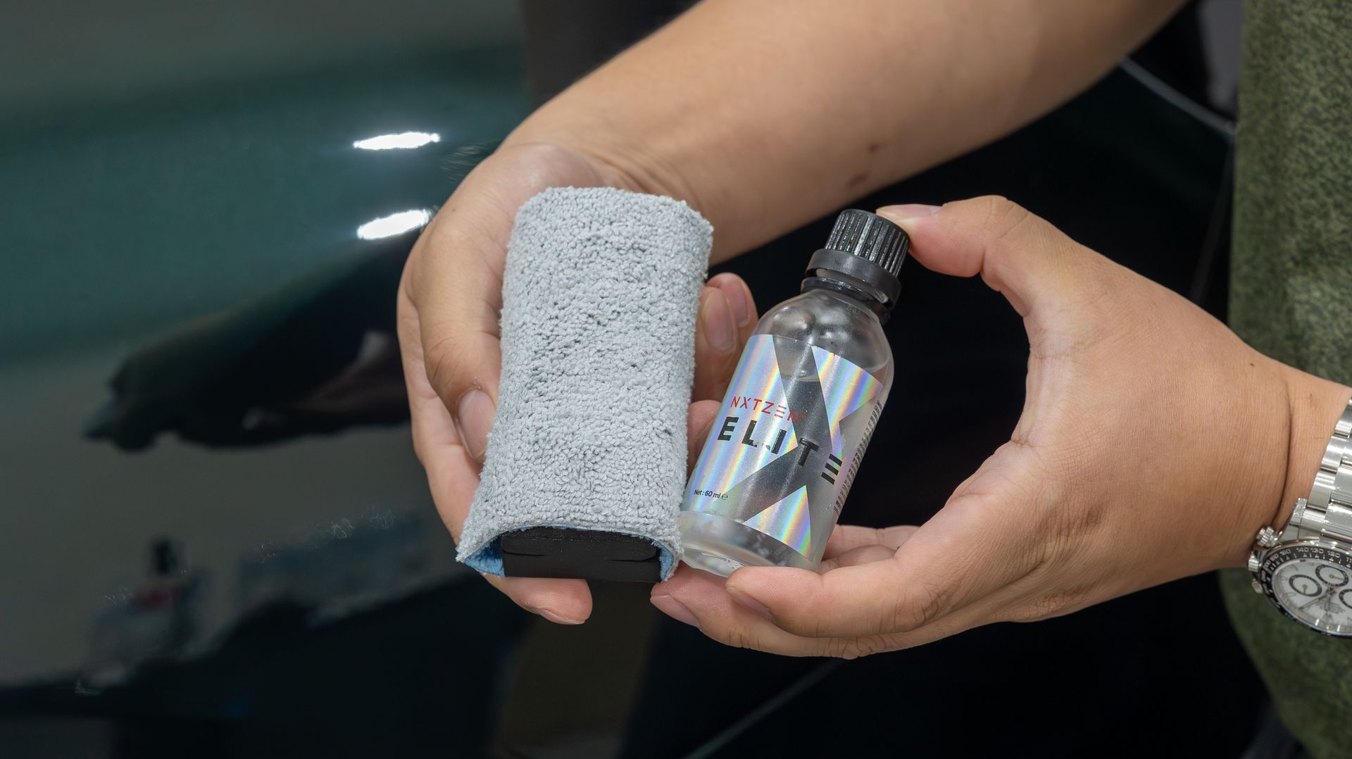 Hands holding a gray foam pad and a clear spray bottle beside a dark car surface.