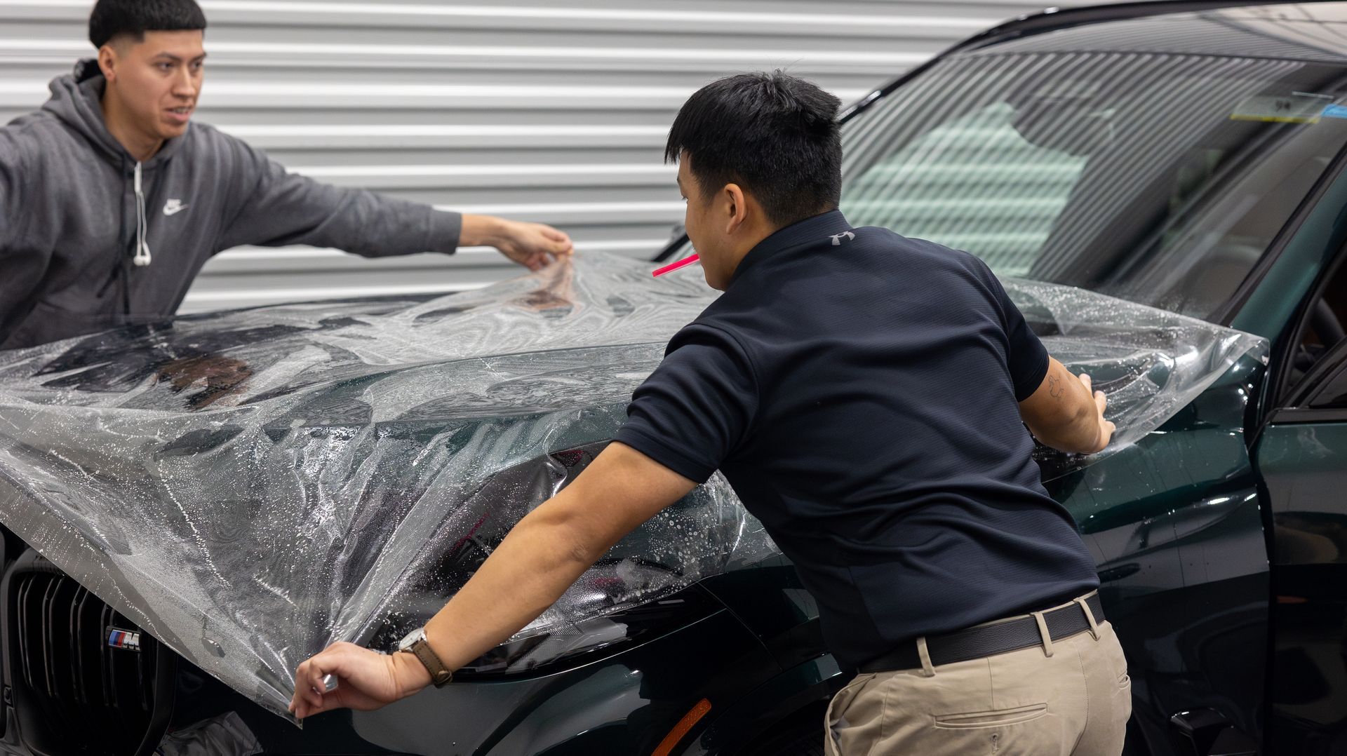Two workers wrapping a car hood with clear protective film in a garage