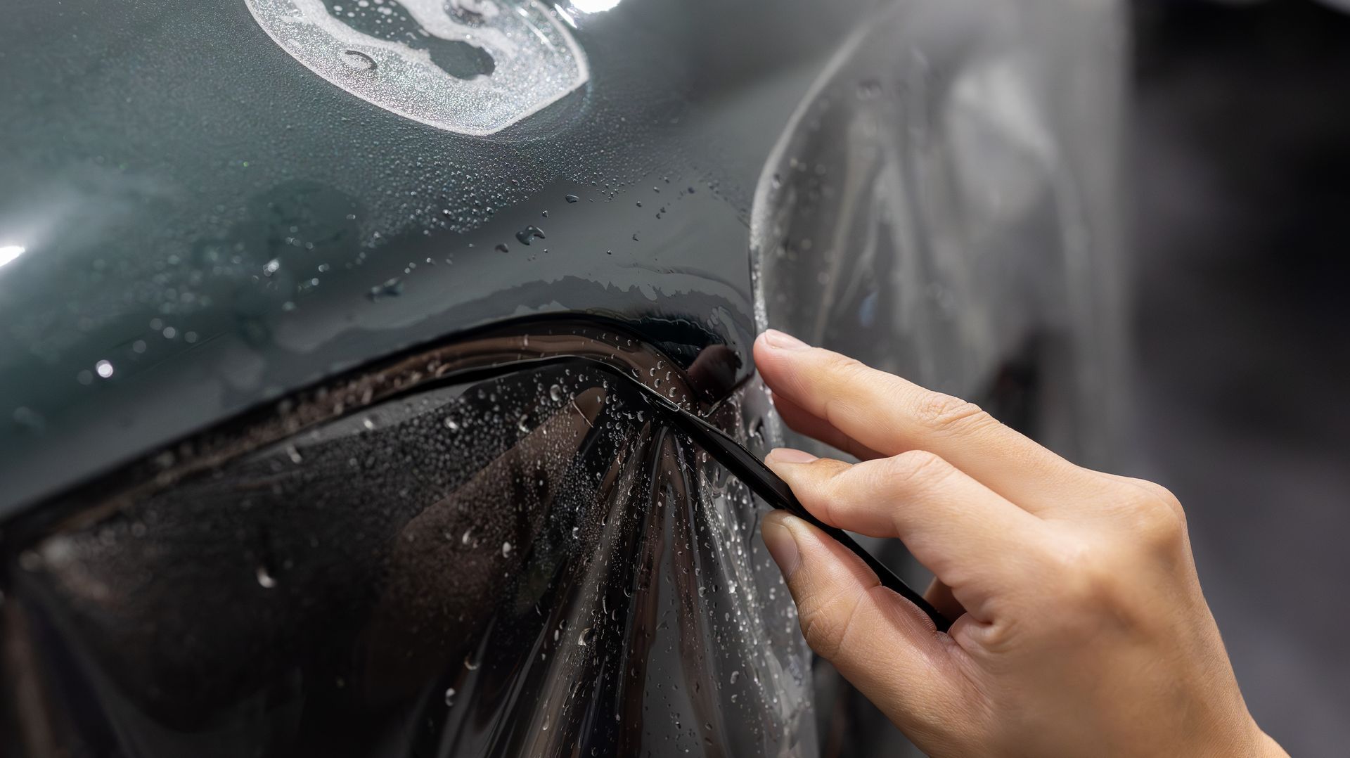 Hand peeling back a black car wrap on a dark green vehicle during detailing