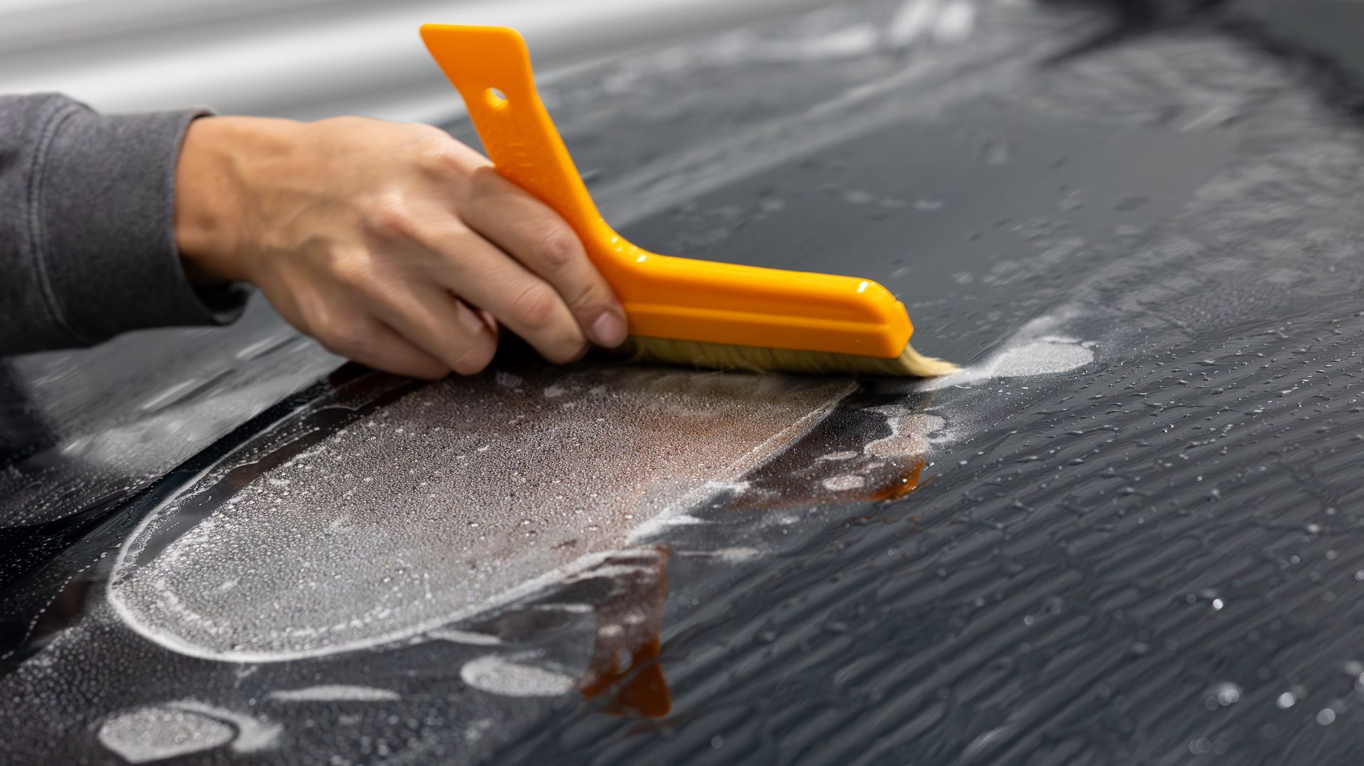 Hand using an orange squeegee to spread repair resin over a crack in a black car windshield.