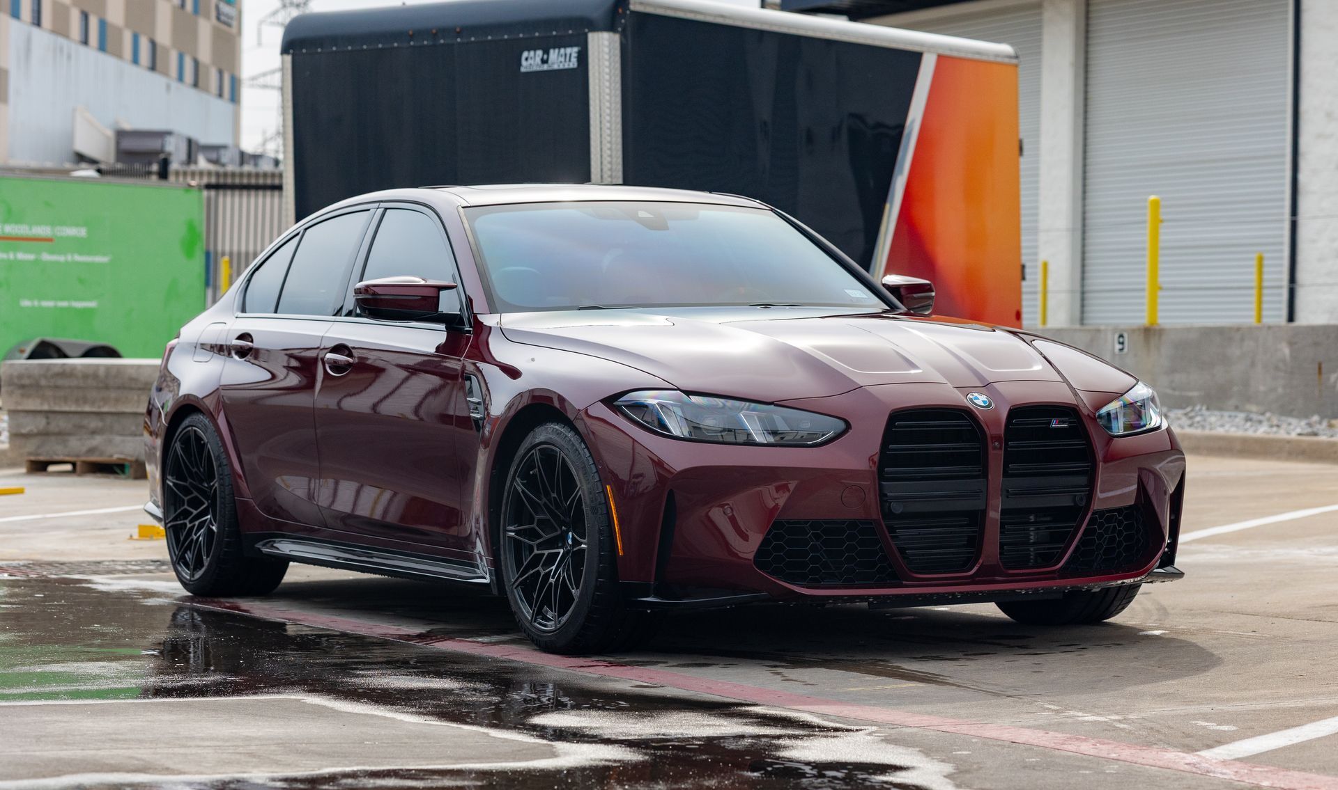 Maroon BMW sedan parked on a wet lot, with black wheels and an open trailer behind it.