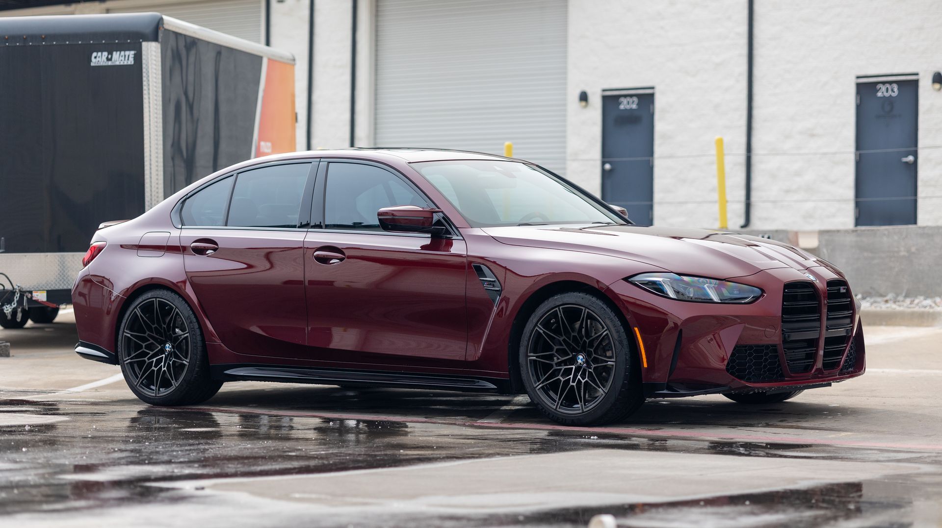 Maroon sports sedan parked on a wet lot beside a warehouse building