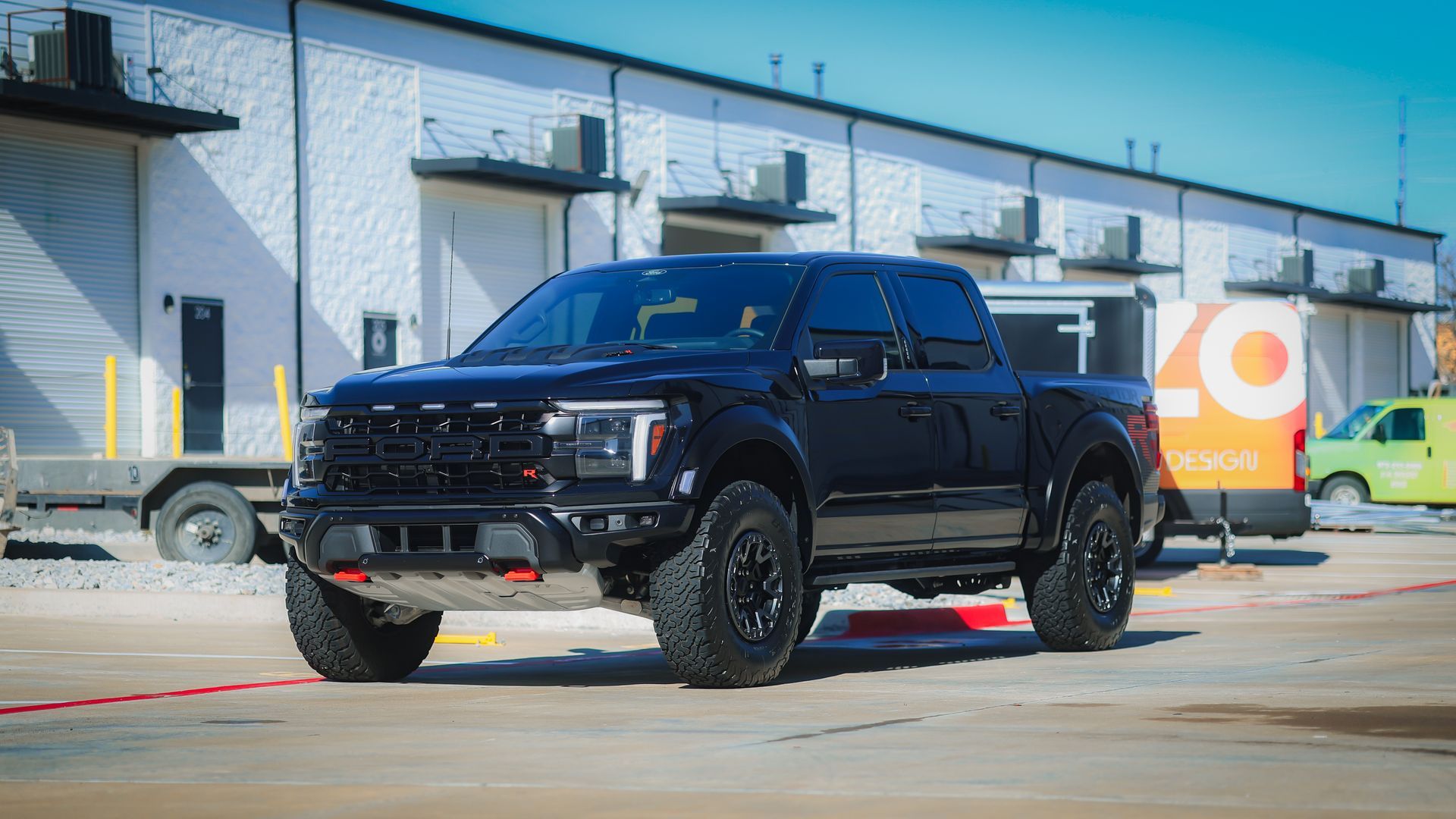 A black Ford F-150 Raptor pickup truck parked in an industrial lot with a warehouse building in the background.